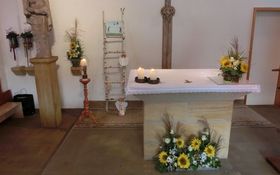 links Marienstatue, daneben brennende Kerze und eine Leiter aus Birkenholz, im Vordergrund ein Altar mit brennenden Kerzen und gelb-weißem Blumenschlmuck