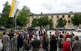 auf einem Kirchplatz stehen viele Leute im Hintergrund weht eine gelb-weiße Fahne