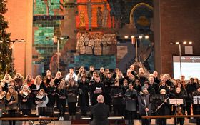 schwarz gekleidete Menschen stehen als Chor aufgestellt, im Hintergrund ein geschmückter Christbaum
