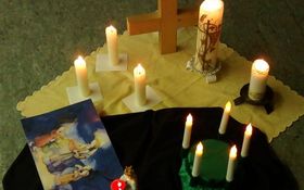 Holzkreuz und diverse brennende Kerzen stehend auf einem gelben Tuch, daneben liegt ein Bild der heiligen Lucia