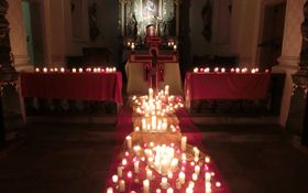 Eine Vielzahl brennender Kerzen auf roten Tüchern, als Kreuzform gelegt. Im Hintergrund steht ein holzkreuz