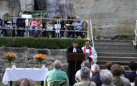 links: Tisch als Altar mit Lilien und Kerzen geschmückt; rechts katholischer und evangelischer Priester im Messgewand, im Hintergrund auf einer Naturbühne ein Posaunenchor