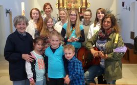 Gruppenfoto mit 10 Kindern und links und rechts je eine erwachsene Frau im Altarraum einer Kirche. Rechte Frau mit umgehngter Gitarre