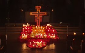 Taizekreuz mit vielen brennenden Kerzen davor, die auf roten und gelben Tüchern stehen