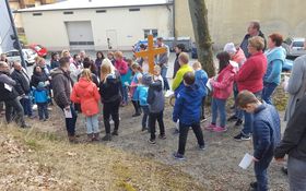 Erwachsene und kinder stehen im Grünen, mittig ragt ein Holzkreuz hervor