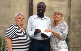 Zwei Frauen überreichen einem dunkelhäutigen Mann ein Kuvert