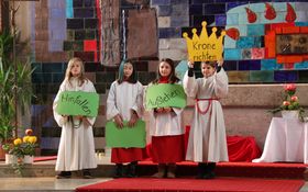 vier Kinder in Ministrantengewändern, eine hält eine Krone aus gelben Karton hoch mit der Aufschrift "Krone richten"