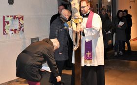 Pfarrer Fenk mit einem Holzkreuz, vor ihm bückt sich eine Frau und legt einen Stein ab