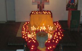 Ein Holzkreuz steht auf einen Tisch, der mit roten und gelben Tüchern bedeckt ist. Die Tücher breiten sich vom Tisch über den Boden aus. Auf den Tüchern stehen brennende Kerzen. Im Vordergrund ist ein Taufstein zu sehen.