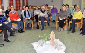 Frauen sitzen in einem Stuhlkreis in der Mitte ein Tuch auf dem Blumen, ein Engel, eine Uhr und ein Stern stehen