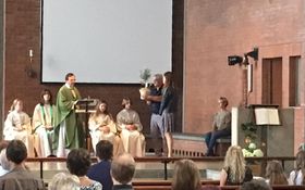 vier Ministranten sitzend, in der Mitte steht ein Pfarrer in grünem Messgewand, rechts daneben stehen ein Mann mit einem Olivenbaum in der Hand und eine Frau