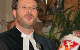 Evangelischer Pfarrer mit Gewand und einer ovalen Kerze in der Hand. Das Motiv der Kerze ist das Dreifaltigkeitssymbol, eine Hand, eine Taube und ein Lamm