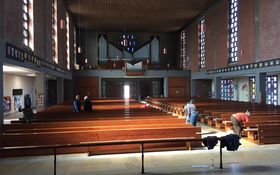 großer Kirchenraum mit vielen leeren Holzbänken, ein paar Erwachsene mit Besen und Lappen stehen im Bild