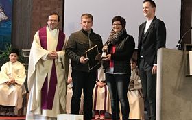 links ein Priester, ein Mann mit Geschenken in den Händen, eine Frau und ein Mann stehen nebeneinander