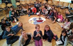 34 Kinder sitzen im Kreis in der Mitte steht ein Kreuz und liegt ein gebasteltes Schiff auf einem blauen Tuch 