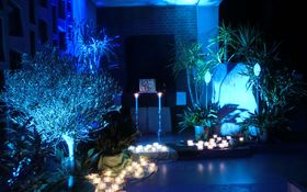 Ein großer beleuchteter Stein umringt mit Palmen steht neben einem Altar, auf dem ein Tabernakel steht. Viele brennende Kerzen in Gläsern davor